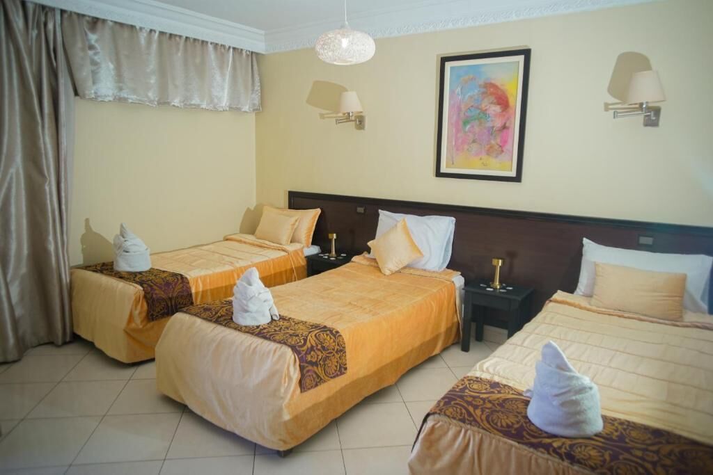 A hotel room with three beds, each with a duvet and pillows, a nightstand, a lamp, and a painting on the wall.