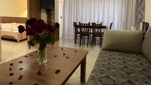 Ein Hotelzimmer mit einer Sitzecke, einem Esstisch mit Stühlen, einem Bett und einer Vase mit roten Blumen.