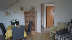 A dining area with a table, chairs, and a vase of flowers. There is a bookshelf with glassware and a door leading to another room. - Wittow Apartment for 2 Guests (50 m²) in Wiek on Rügen (Wiek)