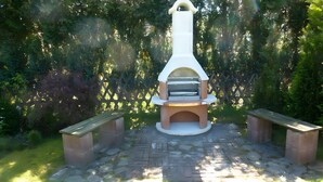 Forno a pizza ao ar livre com chaminé, duas bancadas e área pavimentada em pedra, cercada por vegetação.