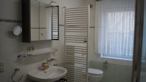 A bathroom with a sink, mirror, towel rack, toilet, and window with blinds. - Modern Apartment for 4 Guests (114 m²) in Wiek on Rügen (Wiek)