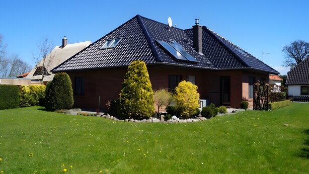 Uma casa com telhado inclinado, painéis solares e um gramado bem cuidado com arbustos e árvores.