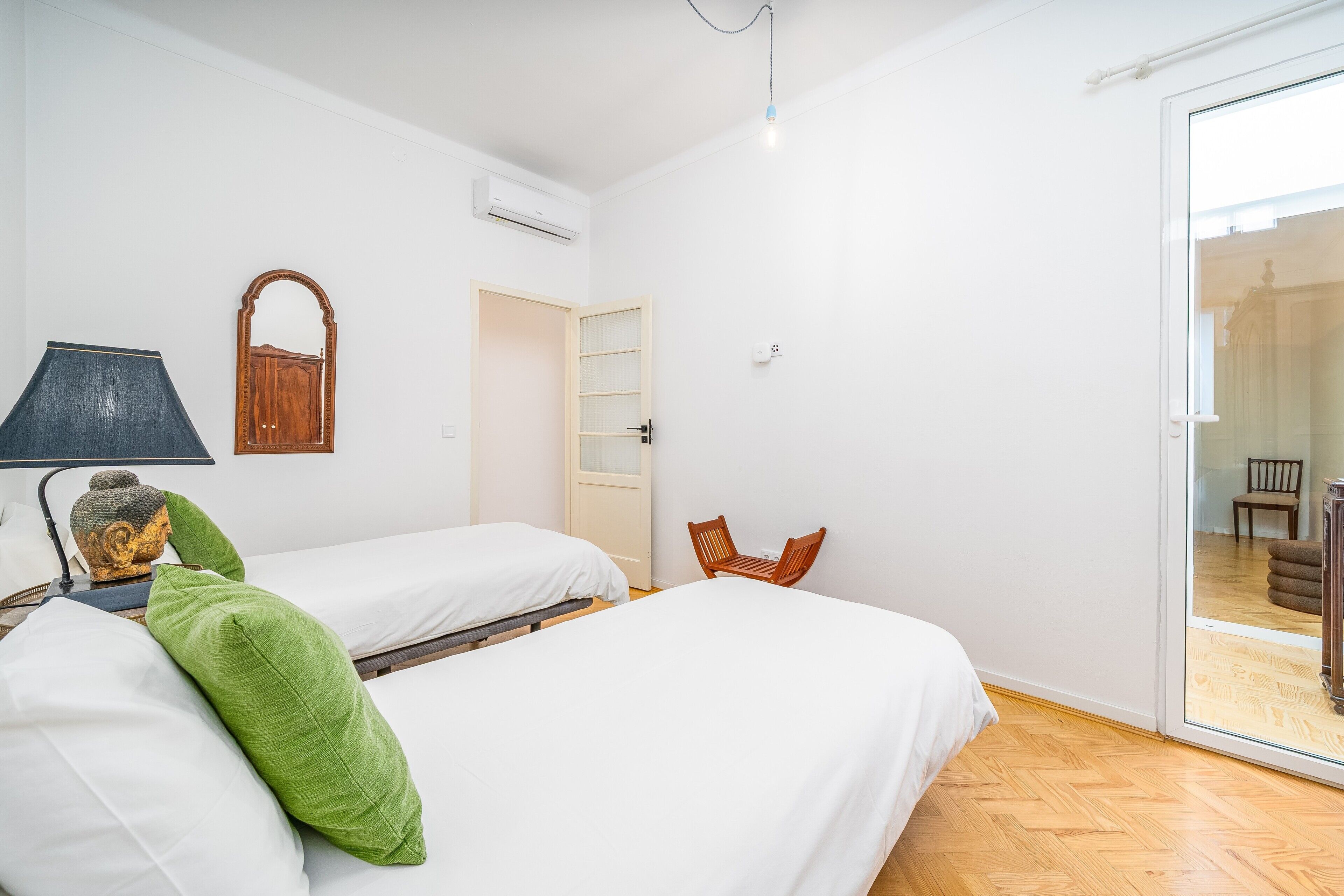 A room with two beds, a chair, a lamp, a mirror, and a sliding glass door to an outdoor area.