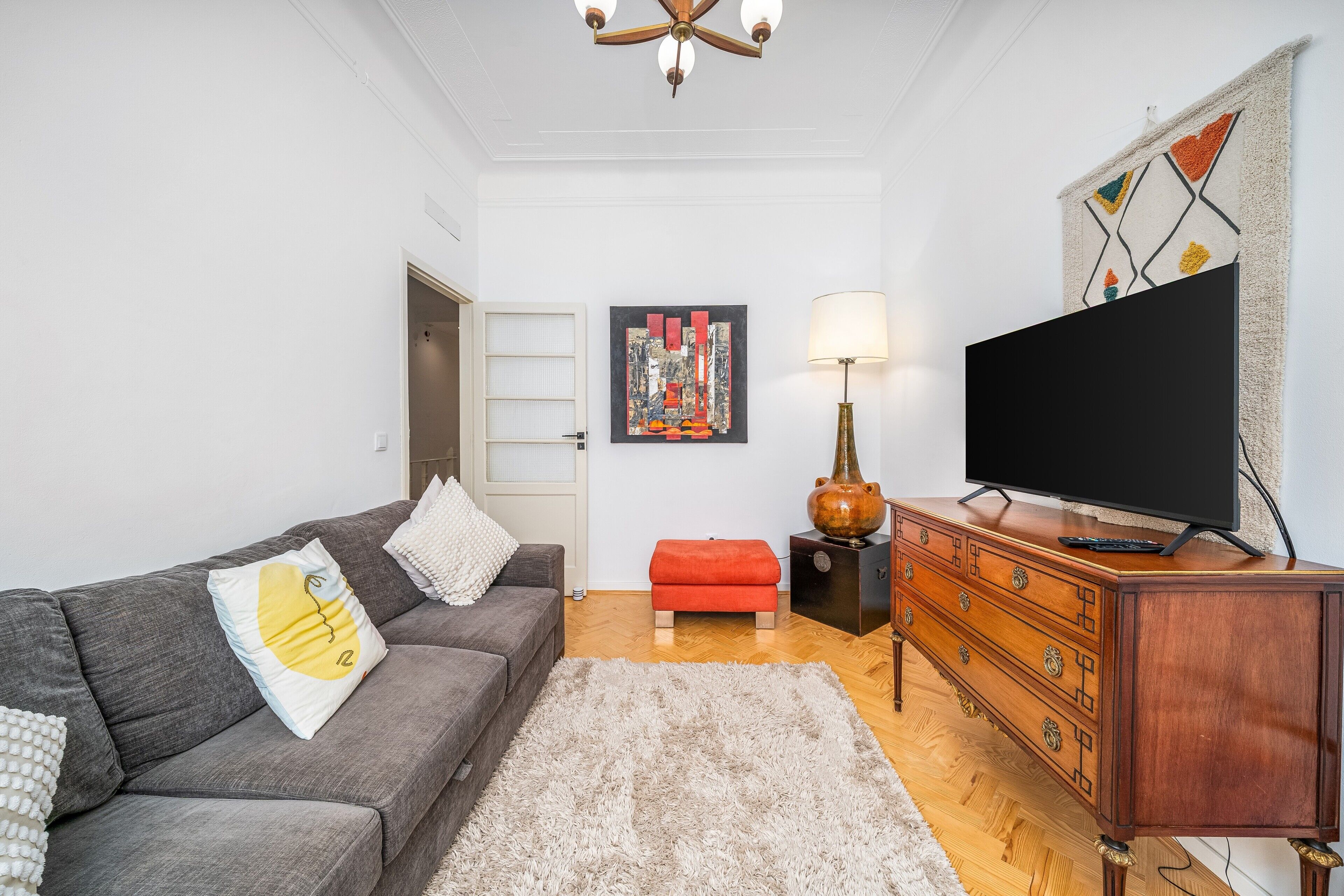 Una sala de estar con un sofá gris, un reposapiés rojo, un mueble de madera, un televisor de pantalla plana y un cuadro en la pared.