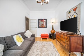Un salon avec un canapé gris, un pouf rouge, un meuble en bois, un téléviseur à écran plat et un tableau accroché au mur.