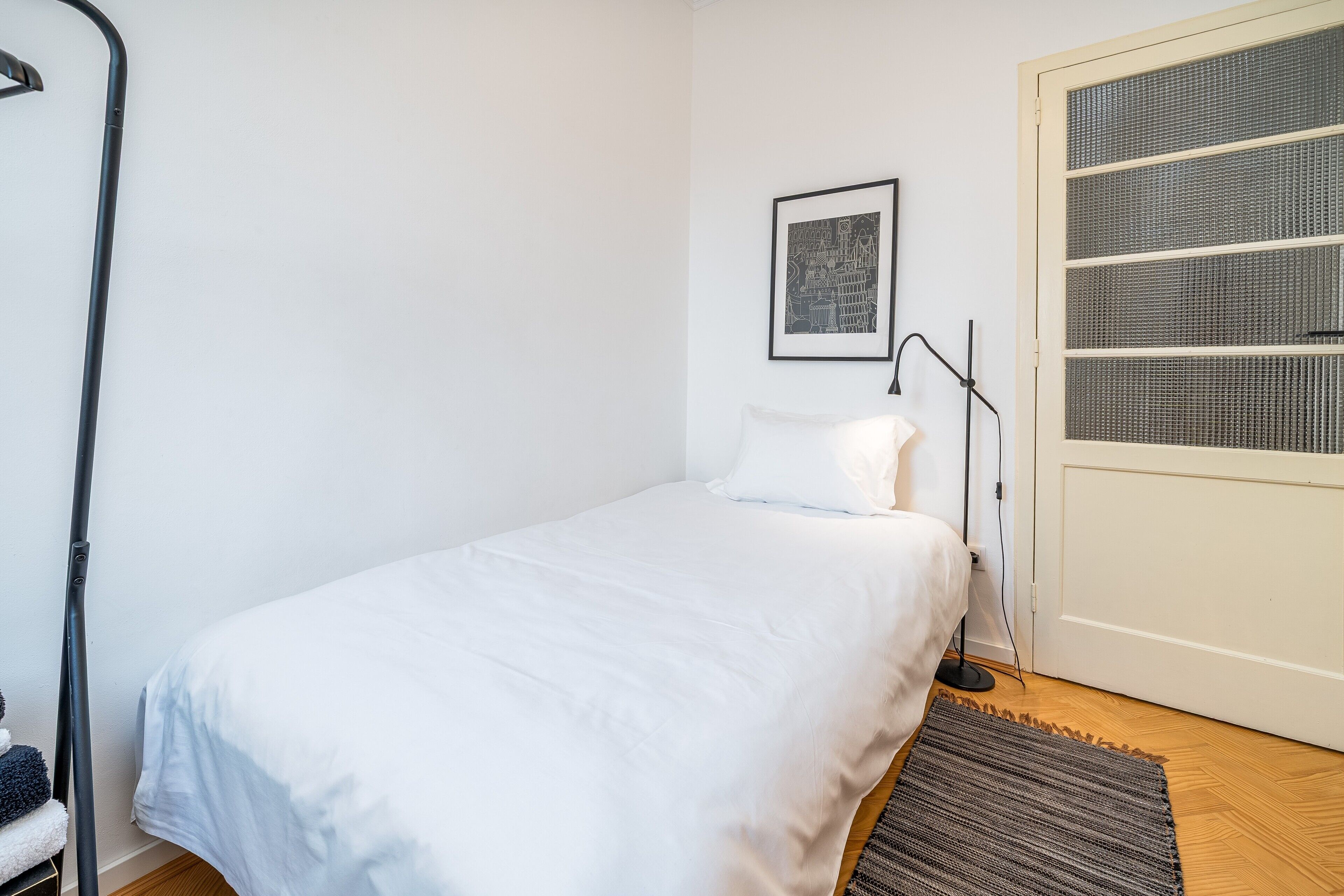 A single bed with white bedding, a framed picture on the wall, a standing lamp, and a door with a mesh screen.