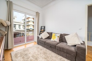 Un salon avec un canapé gris, un tapis moelleux et une vue sur un balcon donnant sur des immeubles.