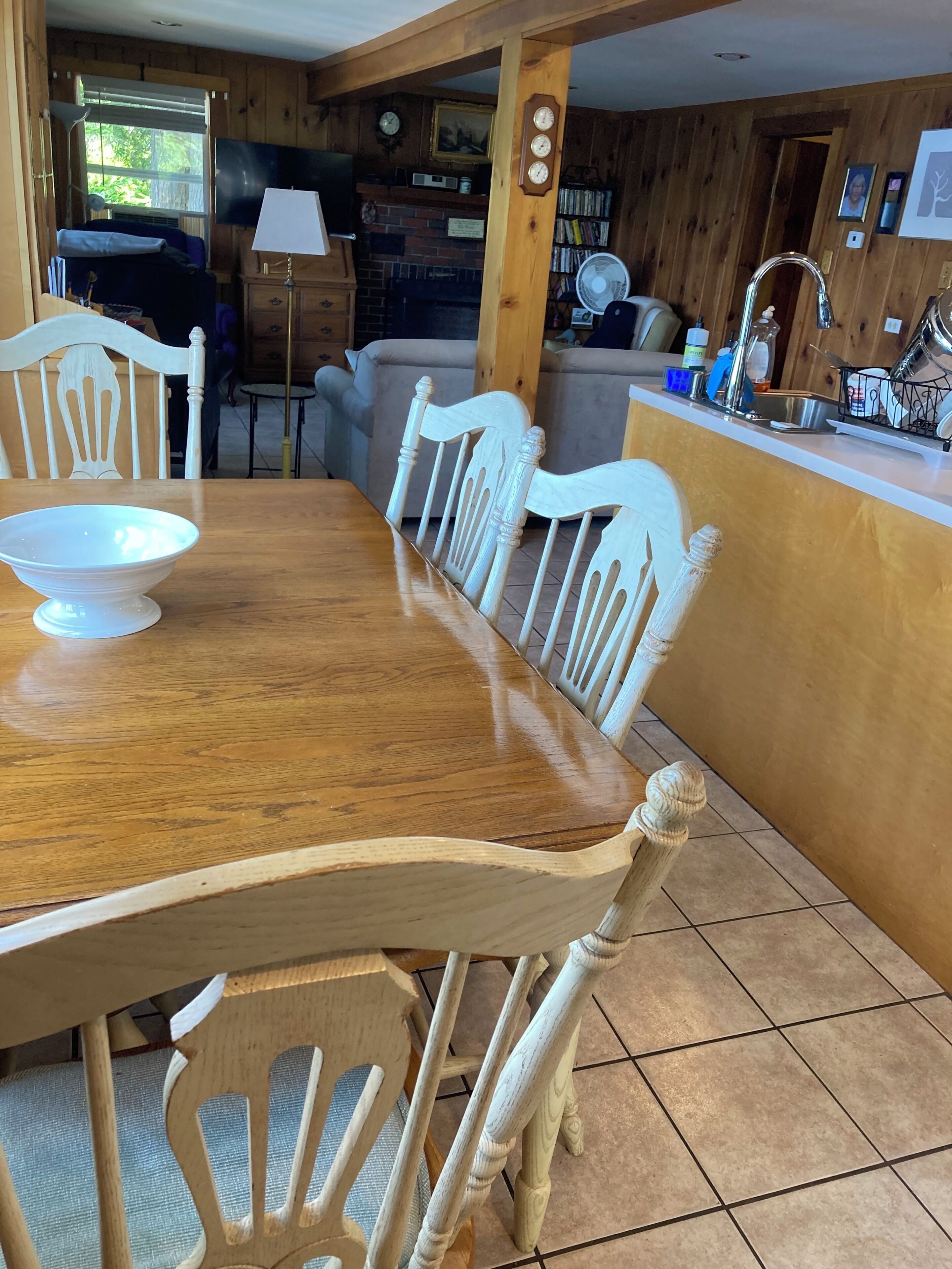 Une table de salle à manger en bois, accompagnée de chaises, un coin cuisine avec un évier et une fenêtre avec des stores.