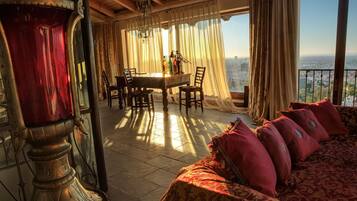A room with a dining table, chairs, a chandelier, a large window with curtains, and a view of a cityscape.