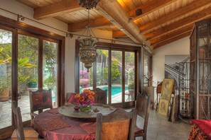 A room with a wooden ceiling, large windows, a round table with a vase of flowers, and a view of a swimming pool.