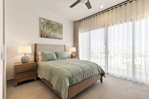 A bedroom with a bed, two bedside tables with lamps, a ceiling fan, a large window with sheer curtains, and a painting on the wall. - 2-bedroom Apt w AC, Gym, Rooftop Infinity Pool Close to Downtown Cabo San Lucas (Cabo San Lucas)