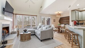 A spacious living room with a fireplace, a grey sofa, a round coffee table, and a kitchen area with wooden stools. - King Bed w Pull Out Sofa + Murphy Bed (Galena)