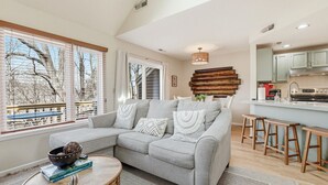 A modern living room with a grey sectional sofa, a small round coffee table, and a kitchen area with a bar stool. - King Bed w Pull Out Sofa + Murphy Bed (Galena)