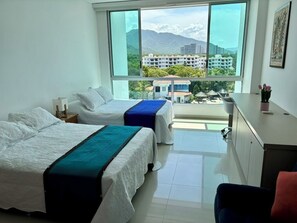 A hotel room with two beds, a desk, and a large window offering a view of buildings and mountains. - Magnificent apartment on the beach of Santa Marta, ideal for families with children. (Santa marta)