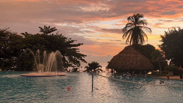 Une piscine agrémentée d’une fontaine, de palmiers et d’une structure surmontée d’un toit de chaume.