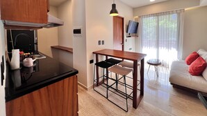 A compact living space with a kitchenette, a dining area with stools, a sofa with cushions, a TV, and a window with curtains. - Tulum & comfort Travel Apartment 2DB 1BR + Jacuzzi, gym, cinema, swimming pools and more (Tulum)