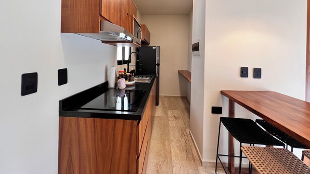 A compact kitchen with wooden cabinets, a black countertop, a built-in stove, and a dining area with a wooden table and black chairs. - Tulum & comfort Travel Apartment 2DB 1BR + Jacuzzi, gym, cinema, swimming pools and more (Tulum)