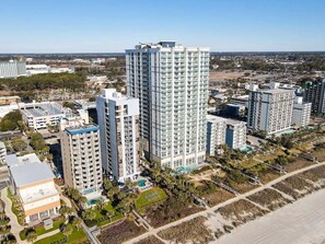 A high-rise building with multiple floors, surrounded by other buildings, a parking area, and a landscaped area with trees. - 2 Bedroom Oceanview Condo - kitchen, W/D, pool, hot tub, gym, racquetball, etc. (Myrtle Beach)