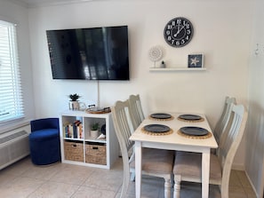 A dining area with a white table, four chairs, a black clock, a TV mounted on the wall, and a blue chair.