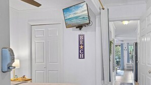 Una camera d'albergo con un letto, una TV fissata al muro, un corridoio che conduce a un balcone e un cartello con la scritta 'BEACH'.