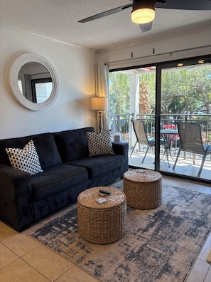 A living room with a dark sofa, two round wicker ottomans, a round mirror, and a ceiling fan.