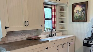 Una cucina con mobili chiari, un lavello, un fornello e una finestra con tenda a quadri bianchi e rossi.