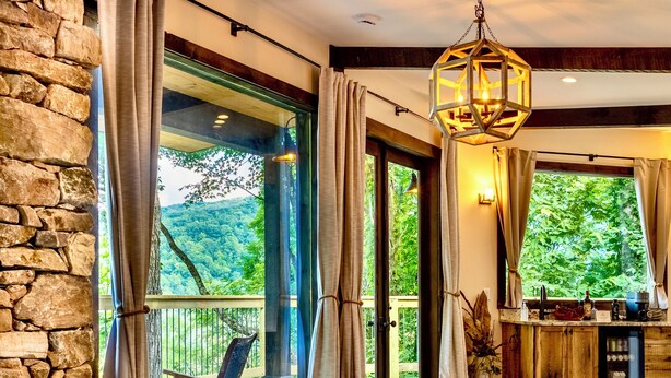 A bedroom with a stone wall, wooden flooring, a large bed, a hanging light fixture, and a view of greenery outside. - Elk & Embers Mountain Hideaway (Waynesville)
