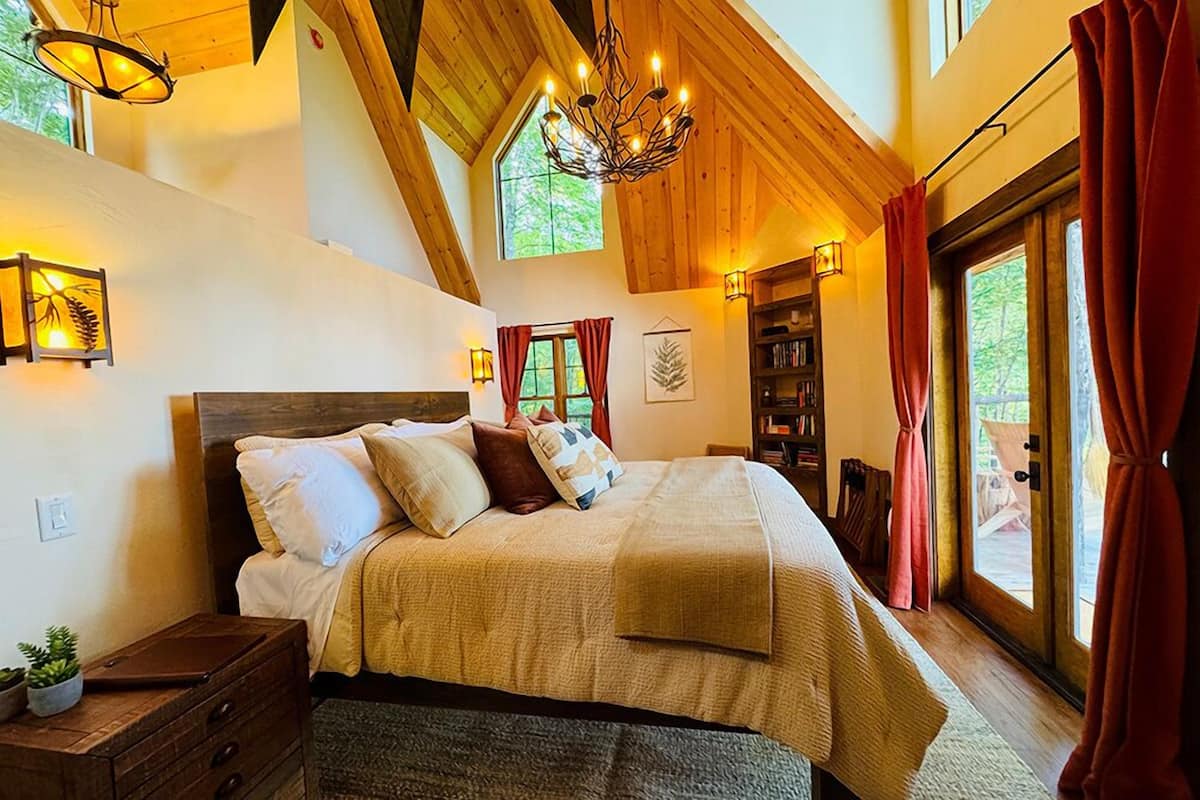 Une chambre avec un grand lit, des poutres apparentes en bois au plafond, un lustre et une vue sur l’extérieur.