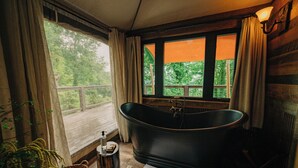 A room with a large bathtub, a window with curtains, a wooden floor, and a small table with a bottle and soap. - Elk & Embers Mountain Hideaway (Waynesville)
