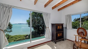 A room with large windows overlooking a body of water, a wooden chair, and a small wooden cabinet. - Te Uenga - Parekura Bay Holiday Home (Parekura Bay)