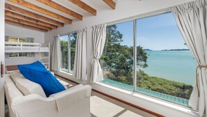 A room with a white sofa, a blue pillow, a bunk bed, and a large window with curtains. - Te Uenga - Parekura Bay Holiday Home (Parekura Bay)