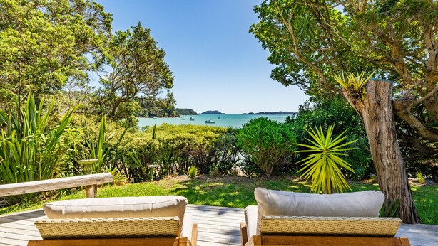 A wooden deck with wicker chairs facing a tropical garden and a view of the sea. - Te Uenga - Parekura Bay Holiday Home (Parekura Bay)
