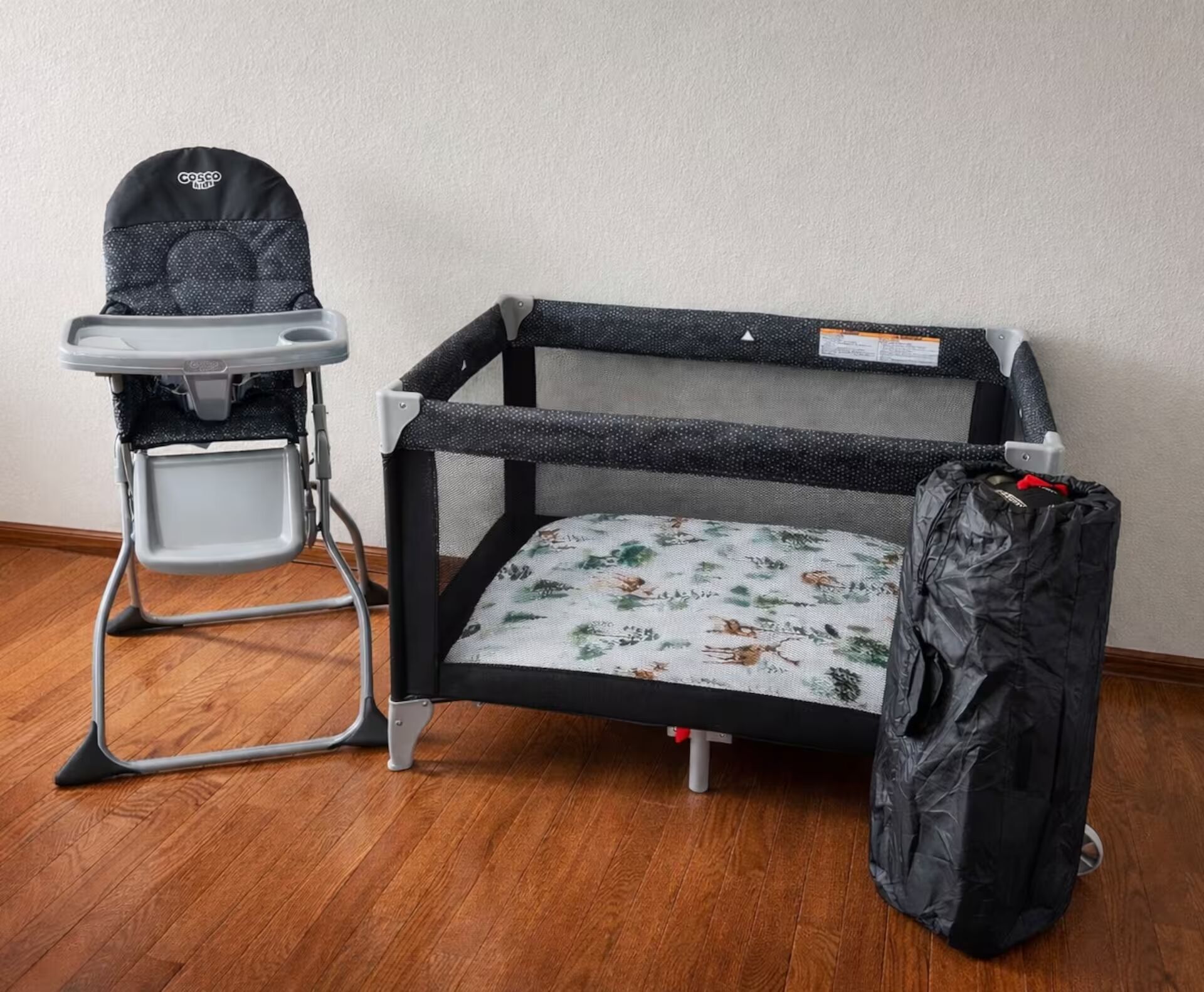 Ein Babybett mit hohem Stuhl daneben, beide mit Netzeinsätzen und grauem Rahmen, auf einem Holzboden vor einer schlichten Wand.