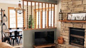 Een woonkamer met een stenen openhaard, een televisie op een houten standaard, een eethoek met tafel en stoelen, en een groot raam met verticale jaloezieën.