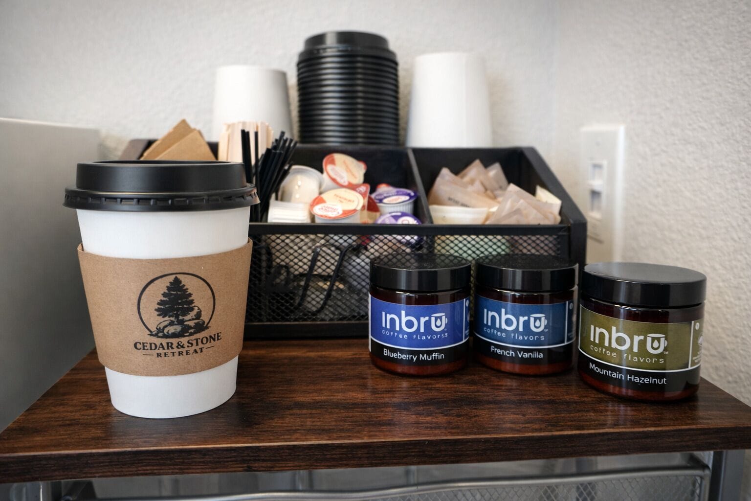 Eine Kaffeetasse mit dem Cedar & Stone Logo, drei Gläser mit Inbru Kaffeearomen und ein schwarzes Tablett mit Kaffeetassen und Besteck.