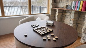 Een ronde houten tafel met een spelbord, een stapel kaarten en verspreide speelstukken. Achtergrond is een boekenplank met boeken, en een raam met uitzicht op een hek en bomen.