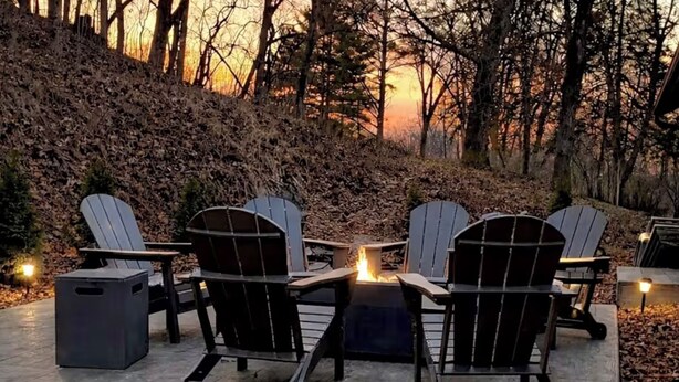 Een vuurkorf omringd door Adirondack-stoelen op een terras, met een bebost heuvel Landschap op de achtergrond.
