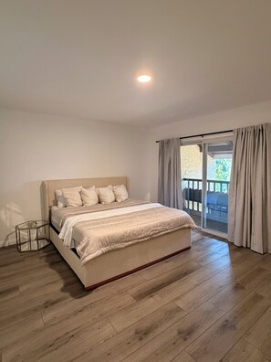 A bedroom with a bed, a glass side table, a wooden floor, and a view of a balcony.