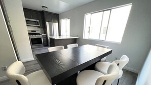 Un coin repas avec une table en bois foncé et des chaises blanches, une cuisine en arrière-plan et une fenêtre laissant entrer la lumière naturelle.