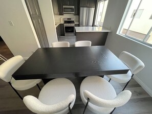 A modern kitchen with stainless steel appliances, a dining area with a black table and white chairs, and a view of the outside through a window.