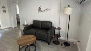 Een woonkamer met een donker leren bank, een houten salontafel, een vloerlamp en een wandairconditioner.