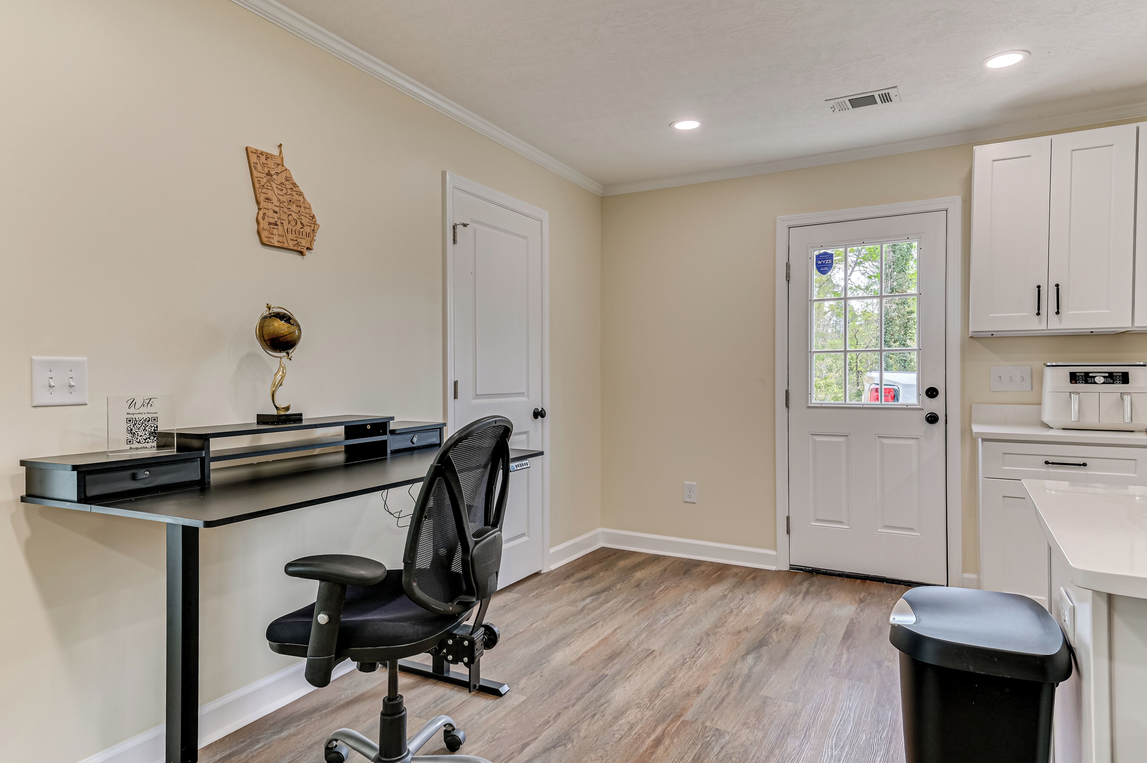 A home office with a desk, chair, and a door leading to a kitchen.