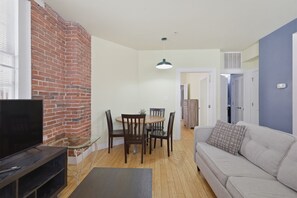 A living room with a TV, a dining table with chairs, a sofa, and a brick wall.