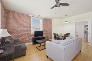 A living room with a brick wall, a television, a sofa, a dining table, and a ceiling fan.