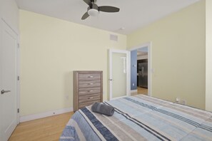 A bedroom with a bed, a dresser, a ceiling fan, and a door leading to a kitchen.