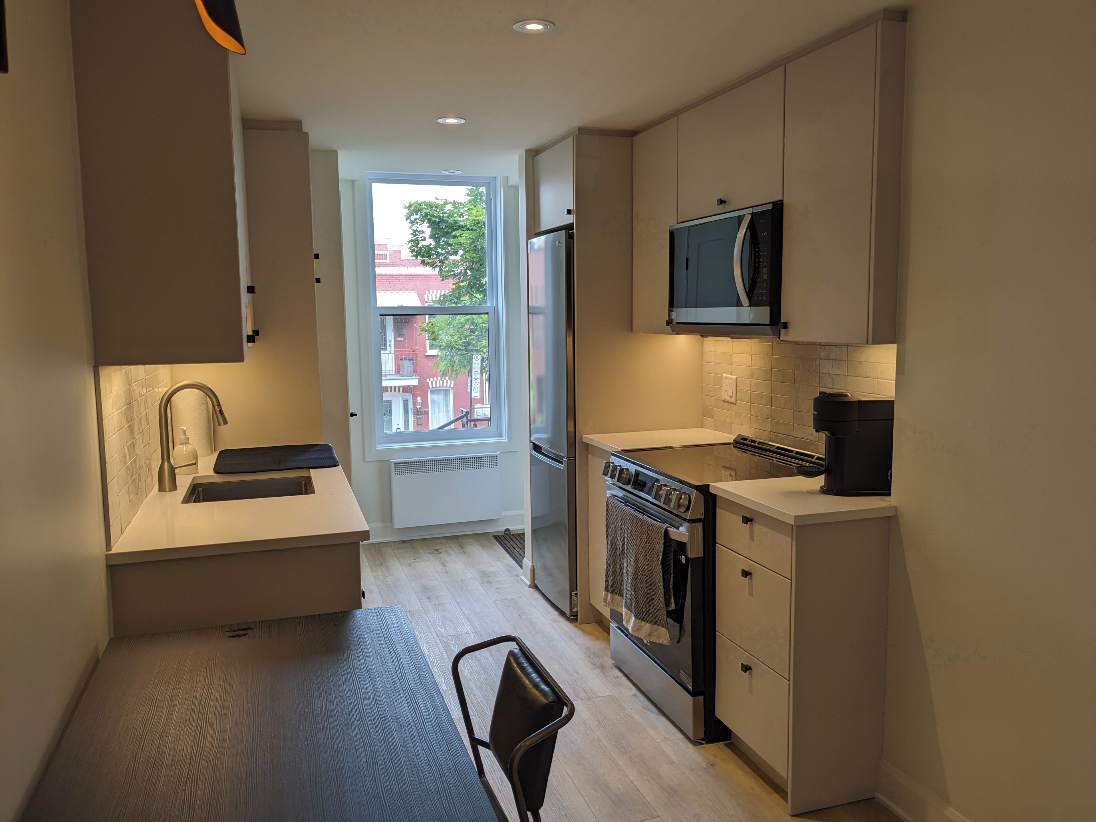 A compact kitchen with a built-in oven, microwave, and sink. There is a chair and a window providing a view of the outside.