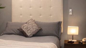 Quarto com cabeceira estofada, colcha cinza, almofada de renda, mesa de cabeceira com abajur e telefone fixado na parede.
