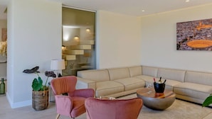 Una sala de estar moderna con un sofá seccional beige, dos sillones rosa, una mesa de centro y un cuadro grande con un paisaje urbano.