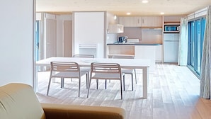 Een moderne woonkamer met een bank, een eettafel met stoelen, een keuken met kasten en apparatuur, en uitzicht door een glazen deur.