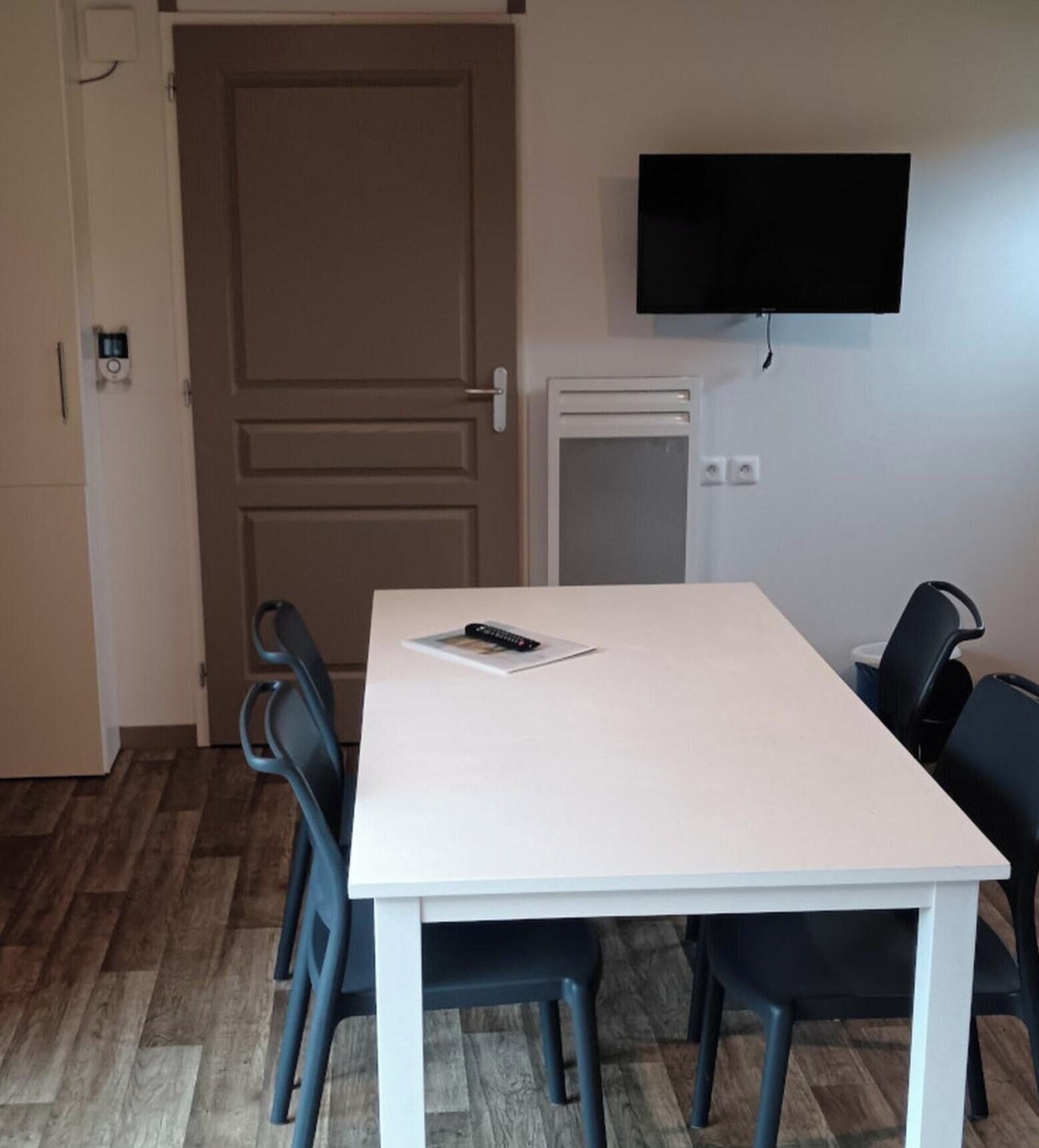 Ett rum med ett vitt bord, fyra stolar, en tv på väggen och en dörr.